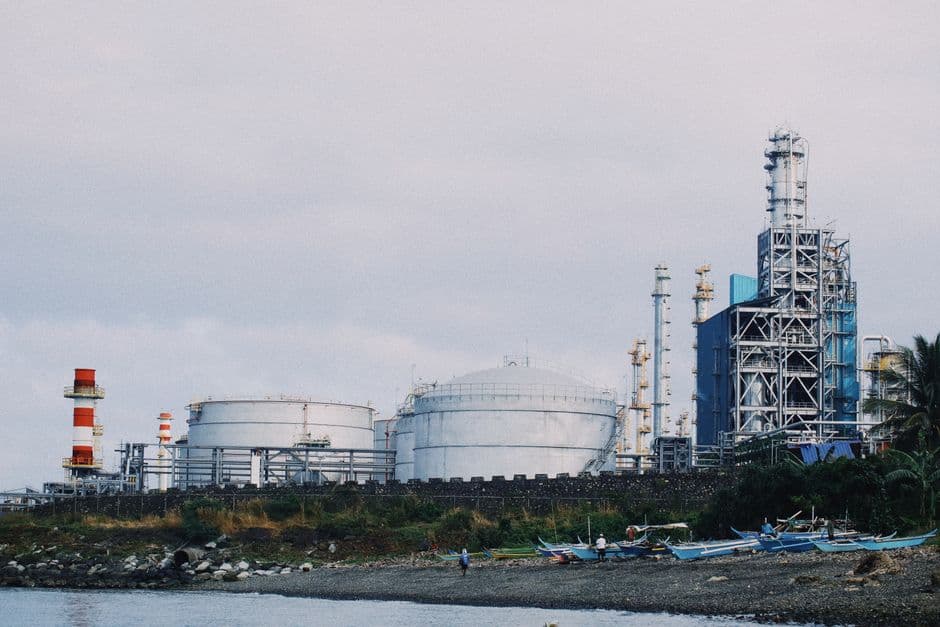 A coastal industrial petrochemical plant with storage tanks and factory structures against a cloudy sky.