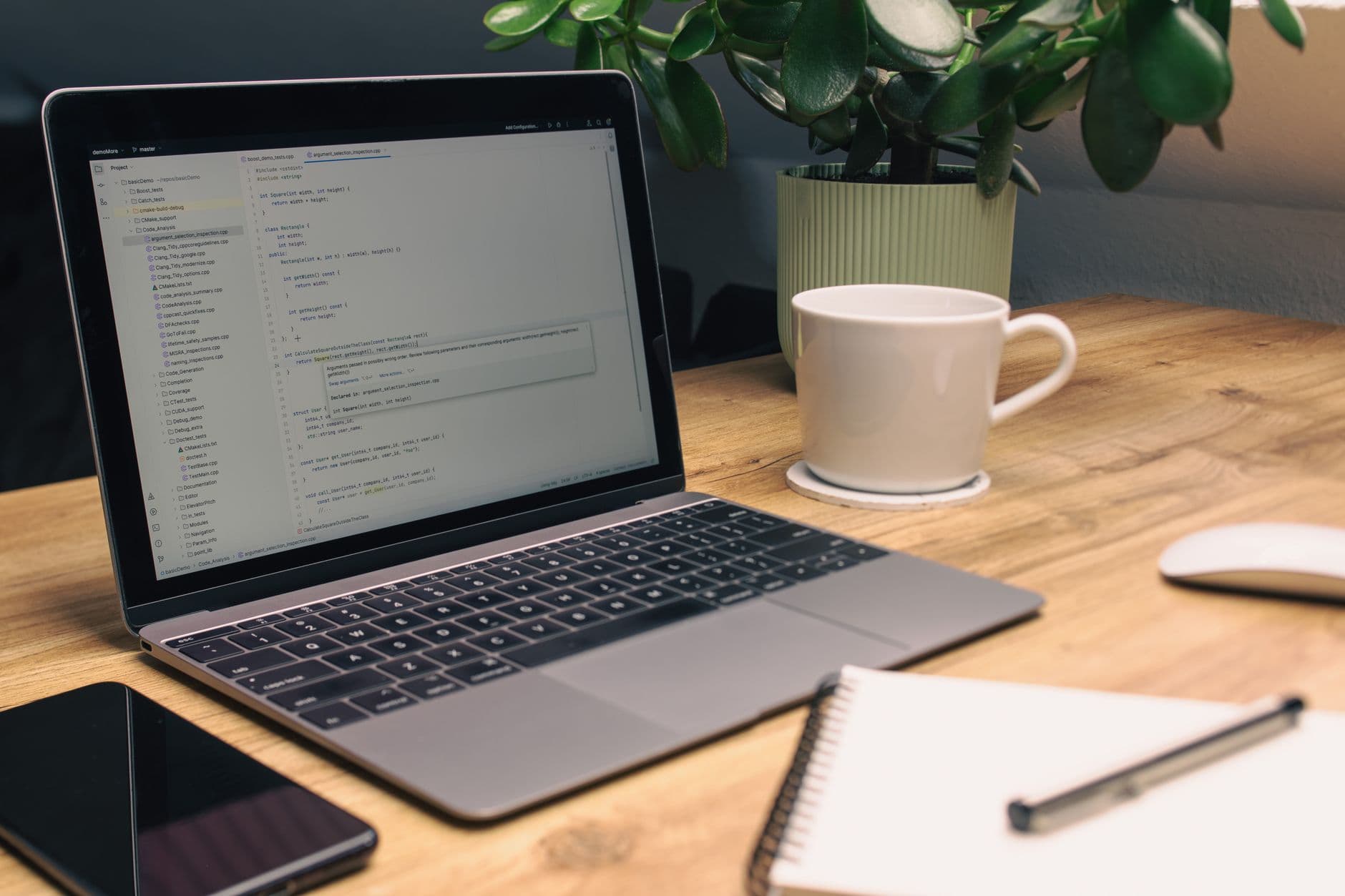 Laptop displaying code on a wooden desk with coffee, plant, and notebook.